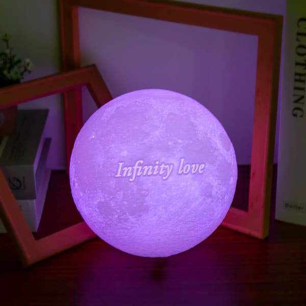 Personalized 3D moon lamp glowing in purple with the engraved text ‘Infinity Love’, placed on soft white fur with decorative stars and rattan balls.
