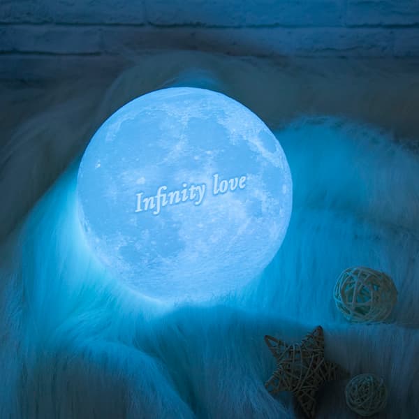 Personalized 3D moon lamp glowing in blue with the engraved text ‘Infinity Love’, placed on soft white fur with decorative stars and rattan balls.