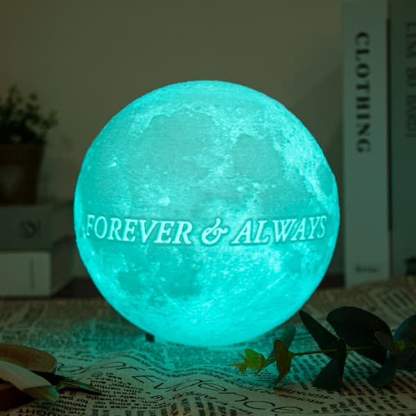 Personalized 3D moon lamp glowing in green with the engraved text ‘Infinity Love’, placed on soft white fur with decorative stars and rattan balls.