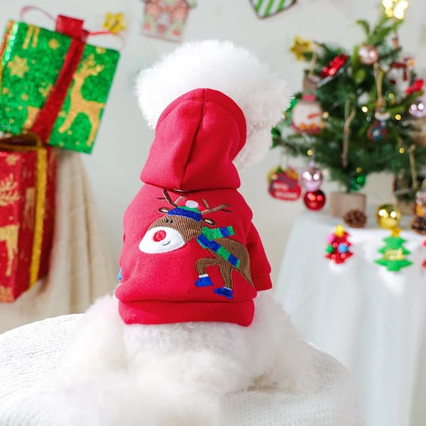 Dog wearing a red hoodie with a reindeer design in a festive setting with Christmas tree and presents.
