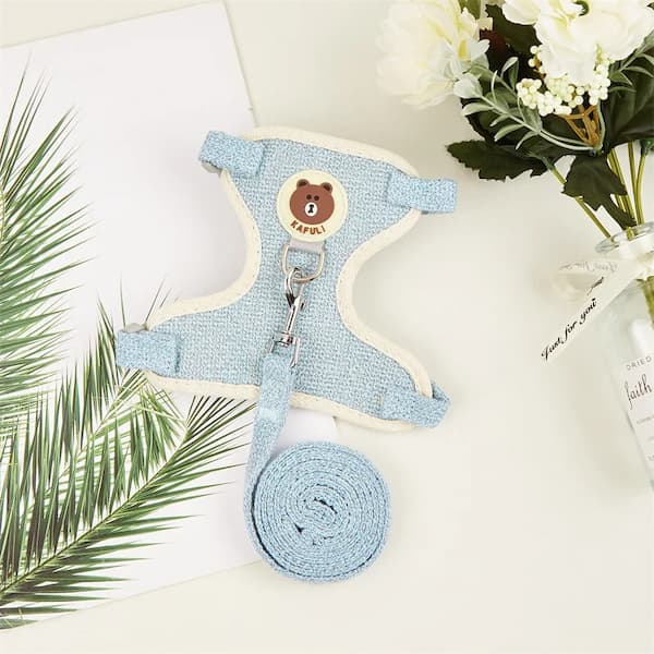 Blue pet harness with a bear design on a white surface with green leaves and a bottle.