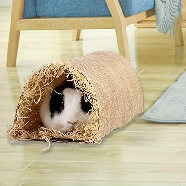Small rabbit peeking out from a straw tunnel on a wooden floor.