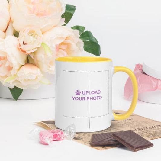 White mug with yellow handle and rim, featuring text 'Upload Your Photo', on a white surface with flowers and chocolate.
