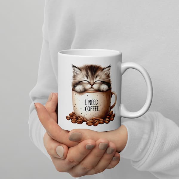 White mug with a cat illustration and 'I Need Coffee' text held by a person against a gray background